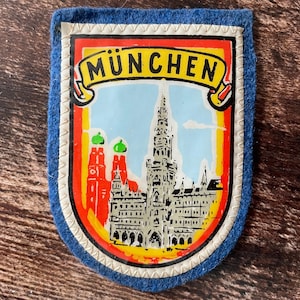 May include: A blue felt patch featuring a graphic illustration of the Munich city skyline, including the Frauenkirche and the New Town Hall. The word MÜNCHEN is printed in black on a yellow banner at the top, framed by a white border with stitching.