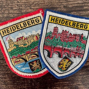 May include: Two shield-shaped embroidered patches featuring the text HEIDELBERG. One patch has a red border, the other a blue border. Both depict a detailed scene of a castle, a bridge over water, and a small crest with a lion emblem in the corner.