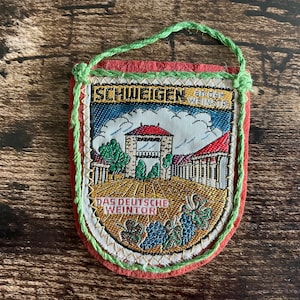 Könnte beinhalten: Ein bestickter Vintage-Souvenir-Wimpel mit dem Text "SCHWEIGEN an der WEINSTRASSE" und "DAS DEUTSCHE WEINTOR". Der Wimpel hat eine rote Rückseite, eine grüne Aufhängeschlaufe und zeigt eine Landschaft mit Gebäuden und Weinreben.