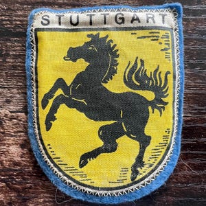 May include: A fabric patch featuring a yellow shield with a black prancing horse silhouette and the text STUTTGART printed in black at the top. The shield is outlined in black and bordered by a blue fabric edge with white stitching.
