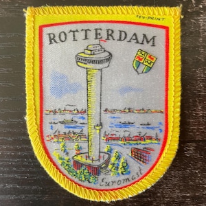 Peut inclure: Un écusson en tissu rectangulaire avec une bordure jaune vif présentant une illustration grise de la tour Euromast à Rotterdam. Le texte ROTTERDAM est imprimé en haut et Euromast en bas. Un petit écusson coloré est également visible.