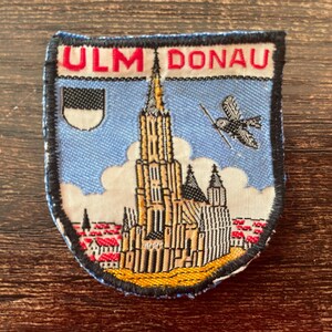 May include: Embroidered patch featuring the words "ULM DONAU" in red. The patch depicts a detailed illustration of a cathedral with a tall spire, a bird, and a coat of arms against a blue sky background. The patch is bordered in black.