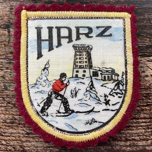 Op de afbeelding: Een schildvormige stoffen embleem met een geschulpte donkerrode vilten rand en een gele gestikte afwerking. Het midden toont een winterse scène met een skiër in een rode jas, een stenen toren en het woord HARZ in zwarte hoofdletters.