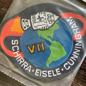 May include: Embroidered patch showing a rocket above a blue globe with green continents. The patch has the text "SCHIRRA • EISELE • CUNNINGHAM" and "VII" in orange. The patch is in a clear plastic sleeve.