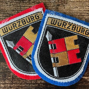 Peut inclure: Deux écussons brodés en forme de bouclier portant le texte WURZBURG. L'un a une bordure en feutre rouge, l'autre une bordure en feutre bleu. Tous deux présentent un fond noir avec un drapeau à damier rouge et or sur une hampe argentée.