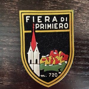 May include: A shield-shaped emblem with the text "FIERA DI PRIMIERO" in white letters. The emblem features a church with a red steeple, a mountain scene, and the number "m. 720". The border is gold, and the background is black.