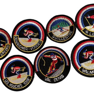 May include: A collection of seven circular embroidered patches featuring skiing scenes. Each patch displays a skier in red and blue clothing on a snowy slope, surrounded by a red, white, and blue border, with text including Trans-Vercors, Gerardmer-Vosges, Guillestre, Les Gets, Les Houches 1008, Val Cenis, and Valmorel.