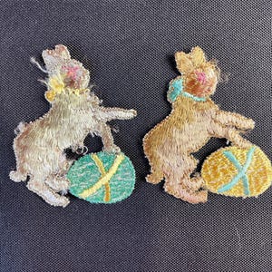 Può includere: Due toppe ricamate di coniglietti pasquali. Un coniglietto è beige con un uovo verde, l'altro marrone con un uovo giallo. Entrambi i coniglietti hanno il naso rosa e tengono in mano un uovo di Pasqua con un disegno a croce.