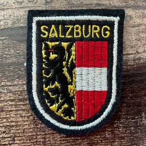 Könnte beinhalten: Ein schwarzer gestickter Aufnäher mit dem Text SALZBURG in gelbem Garn. Das schildförmige Design zeigt links einen schwarzen Löwen auf gelbem Grund und rechts horizontale rote und weiße Streifen, alles umrandet von einer weißen Naht.