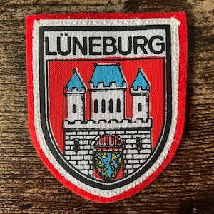 May include: A shield-shaped patch with a red border and a white and black trim. The patch displays the word "LÜNEBURG" above a castle graphic with blue turrets on a red background.