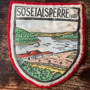 Könnte beinhalten: Ein schildförmiger Stoffaufnäher mit rotem Rand, der eine gedruckte Illustration der Sösetalsperre Harz zeigt. Das Motiv stellt einen See, grüne Hügel, ein Gebäude mit rotem Dach und eine Straße in einem klassischen, illustrativen Stil dar.