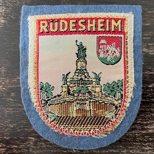May include: A rectangular blue felt patch featuring a woven design of the Niederwald Monument in Rudesheim. The patch includes the text RÜDESHEIM in black letters on a red background, a small crest, and a gold-coloured border with white stitching.
