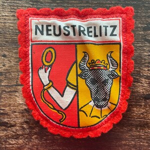 May include: A fabric patch featuring the coat of arms of Neustrelitz with a scalloped red felt border. The shield is divided into red and yellow sections, displaying a white arm holding a gold ring and a black bull head wearing a crown.