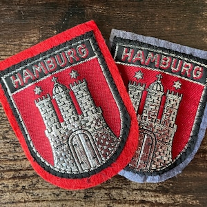 May include: Two embroidered patches with the word "HAMBURG" above a silver castle illustration. One patch has a red background with black trim, the other a grey background. The patches are shield-shaped.