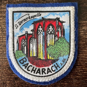 May include: A shield-shaped fabric patch with a blue felt border. The centre features a graphic illustration of the St. Wernerkapelle church in red, yellow, and white against a blue sky. The text reads St. Wernerkapelle and BACHARACH A.RH.