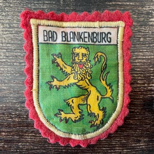 May include: A rectangular patch with a red scalloped border and a green center. The patch features a yellow lion emblem and the text "BAD BLANKENBURG" in black. The patch is likely a souvenir or emblem.