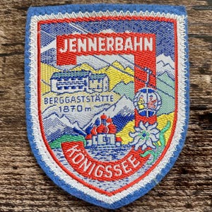 Könnte beinhalten: Ein schildförmiger Stickaufnäher mit einer Berglandschaft und dem Text JENNERBAHN, BERGGASTSTÄTTE 1870 m sowie KÖNIGSSEE. Das Design zeigt eine Seilbahn, ein Gebäude, eine Kirche mit roten Kuppeln und eine Edelweißblüte in Blau und Rot.