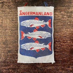 May include: A rectangular patch with the word "ÅNGERMANLAND" in red at the top. Below, three white fish with red fins are displayed on a blue background. The patch has a white border and is made of fabric.