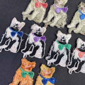 Può includere: Toppe ricamate a forma di gatto in vari colori. Le toppe raffigurano gatti con papillon nei toni del rosso, viola, verde e blu. I gatti sono nei toni del beige, arancione, nero e bianco. Le toppe sono su uno sfondo scuro.