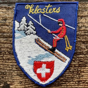 Op de afbeelding: Een schildvormige geborduurde patch met een blauwe achtergrond en de gele tekst Klosters. Het ontwerp toont een skiër in een rood pak op een witte besneeuwde helling met grijze bomen en een klein rood schild met een wit kruis onderaan.