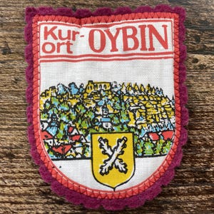 Op de afbeelding: Een rechthoekige stoffen patch met een geschulpte, donker magenta vilt rand. Het witte midden bevat de rode tekst Kur-ort OYBIN boven een kleurrijke illustratie van een stadslandschap en een geel schild met twee gekruiste witte takken.