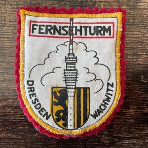 May include: A fabric patch with a red, yellow, and white design. The patch features the text "FERNSEHTURM" at the top, a drawing of a tower, and the words "DRESDEN" and "WACHWITZ" on the sides. The patch has a decorative red border.
