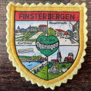 May include: A shield-shaped fabric patch with a scalloped yellow felt border. The patch features a red header with the text FINSTERBERGEN and four illustrated quadrants depicting local scenes, with a central green heart labelled Gruß aus Thüringen.