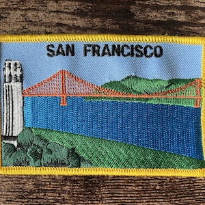 Pode incluir: Um emblema retangular bordado com um céu azul-claro e o texto SAN FRANCISCO em preto. O design inclui um farol branco, uma ponte suspensa laranja brilhante, um corpo de água azul-escuro e colinas verdes.