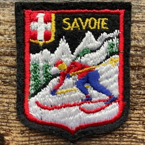 Op de afbeelding: Een schildvormige geborduurde patch met een skiër in een rood jack en blauwe broek op een besneeuwde berg. Het woord SAVOIE is in het geel bovenaan geborduurd, met een klein rood-wit kruisembleem in de linkerbovenhoek, alles omrand in rood.