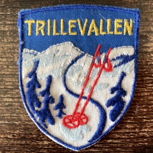Op de afbeelding: Een schildvormige geborduurde patch met de tekst TRILLEVALLEN in geel garen op een blauwe achtergrond. Het ontwerp toont een wit besneeuwd berglandschap met donkerblauwe dennenbomen en twee felrode skistokken met sneeuwtellers.