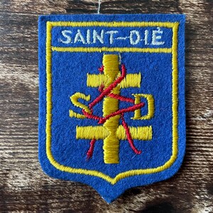 Peut inclure: Un écusson en feutrine bleue avec une bordure jaune et le texte SAINT-DIÉ en blanc. Le centre présente une croix à double traverse jaune avec les lettres S et D en jaune de chaque côté, le tout recouvert d'un fil rouge brodé enchevêtré.