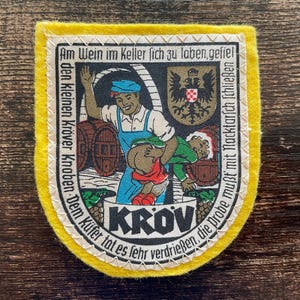 Könnte beinhalten: Ein gelber Vintage-Patch mit einer genähten Bordüre. Der Patch zeigt eine farbenfrohe Illustration eines Mannes und eines Kindes in der Nähe von Fässern mit deutschem Text. Das Wort "KROV" ist deutlich sichtbar.