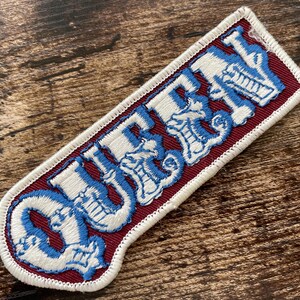 May include: A rectangular embroidered patch featuring the word QUEEN in white block letters with a blue outline. The text is set against a deep maroon background, and the entire patch is finished with a thick white stitched border.