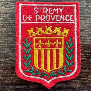 May include: A rectangular red felt patch featuring white embroidered text that reads ST REMY DE PROVENCE. Below the text is a yellow embroidered mural crown above a shield with three yellow fleur-de-lis and vertical stripes, flanked by green laurel branches.
