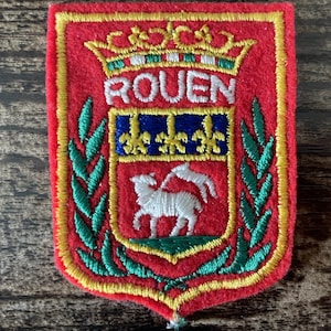 Pode incluir: Um emblema retangular de feltro vermelho com o brasão de armas de Rouen bordado. O design inclui uma coroa amarela acima do texto ROUEN, uma seção azul com três flores-de-lis amarelas e um cordeiro branco com uma bandeira em fundo vermelho.