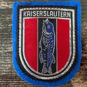 May include: A blue, embroidered patch with the word "KAISERSLAUTERN" at the top. The patch features a red and black shield design with a blue fish in the center. The patch has a silver border.