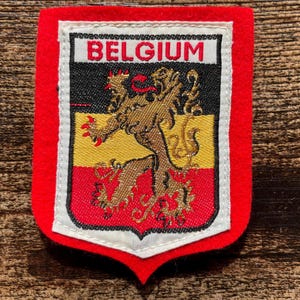 Può includere: Una toppa ricamata a forma di scudo con un bordo in feltro rosso e un emblema centrale con la scritta BELGIUM in rosso. L'emblema raffigura un leone dorato su uno sfondo a strisce orizzontali nere, gialle e rosse con finiture bianche.