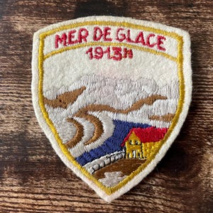 Könnte beinhalten: Gestickter Aufnäher mit dem Text "MER DE GLACE 1913m" in Rot. Der Aufnäher zeigt eine Berglandschaft mit einem Haus, einem Fluss und schneebedeckten Bergen. Der Aufnäher hat einen gelben Rand und einen cremefarbenen Hintergrund.