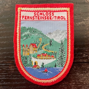 Könnte beinhalten: Ein rechteckiger gestickter Aufnäher mit rotem Filzrand, der eine Landschaftsszene von Schloss Fernsteinsee in Tirol zeigt. Das Design umfasst ein gelbes Schloss, grüne Berge, einen blauen See mit einem kleinen Boot und den Text Schloss Fernsteinsee/Tirol.