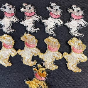 Può includere: Toppe ricamate a forma di orsetto in bianco e oro, con colletti rosa e contorni neri. Le toppe sono disposte su uno sfondo scuro, mostrando le cuciture dettagliate e il design delle figure degli orsetti.