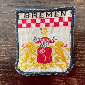 Può includere: Toppa rettangolare con la scritta "BREMEN" in bianco sopra un motivo a scacchi rossi e bianchi. Sotto, uno stemma con uno scudo rosso, leoni dorati e dettagli ornati.