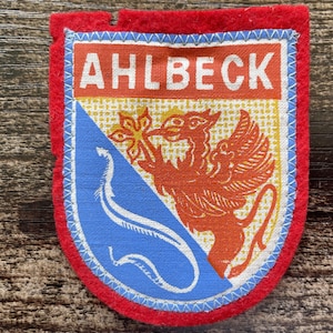 May include: A shield-shaped fabric patch with a red felt border. The center features the text AHLBECK in white on an orange background. Below, a blue diagonal section contains a white curved line, and a red griffin stands on a yellow grid background.