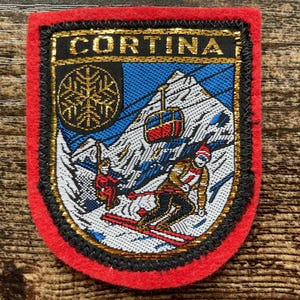 May include: A rectangular embroidered patch with a red felt border featuring the text CORTINA in gold. The design depicts a snowy mountain landscape with a ski lift, a cable car, a snowflake emblem, and a skier wearing a red hat and jacket.