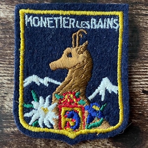 Op de afbeelding: Een rechthoekige donkerblauwe geborduurde patch met een gele rand. Het bevat de witte tekst MONETIER LES BAINS bovenaan, een bruine steenbokkop in het midden, witte bergtoppen, een witte edelweissbloem en een klein kleurrijk wapenschild.