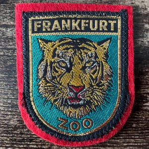 Könnte beinhalten: Ein rechteckiger gestickter Aufnäher mit rotem Filzrand. Die Mitte zeigt ein türkisfarbenes Schild mit einer goldenen Tigerkopf-Illustration. Der Text FRANKFURT ist oben in Gold gestickt, und das Wort ZOO ist unten in Gold gestickt.