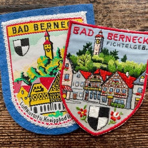 May include: Two embroidered fabric patches featuring the town of Bad Berneck. The left patch has a blue felt border and yellow text. The right patch has a red border and depicts a town scene with a clock tower, houses, and a black and white shield.