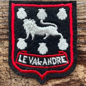 Puede incluir: Un parche bordado en forma de escudo con fondo de fieltro negro y borde rojo. El diseño incluye una figura de león blanco rodeada por cinco formas de concha de vieira blancas. Una cinta roja en la parte inferior contiene el texto blanco LE VAL-ANDRE.