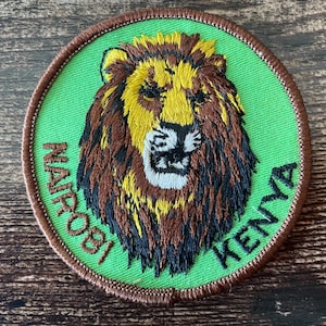 Può includere: Toppa rotonda ricamata con la testa di un leone con criniera gialla e pelliccia marrone su sfondo verde. La toppa è bordata con cuciture marroni e riporta le parole "Nairobi Kenya".