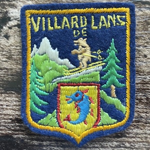Op de afbeelding: Een rechthoekige geborduurde patch met een donkerblauwe viltachtergrond en een gele rand. Het bevat de tekst VILLARD LANS DE, een beige skiër, witte bergtoppen, groene dennenbomen en een geel schild met een blauwe dolfijnafbeelding.