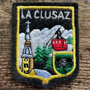 May include: A rectangular embroidered patch with a black background and a yellow border. It features the white text LA CLUSAZ above a scene with a church steeple, green pine trees, white snow-capped mountains, a red cable car, and a green shield.